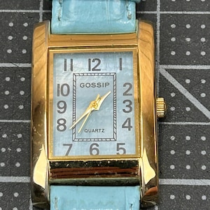 May include: A rectangular gold-coloured wristwatch with a light blue face and matching strap. The watch face displays the word "GOSSIP" and the word "QUARTZ". The watch has gold-coloured hands and hour markers. The strap is a light blue, textured leather-like material.