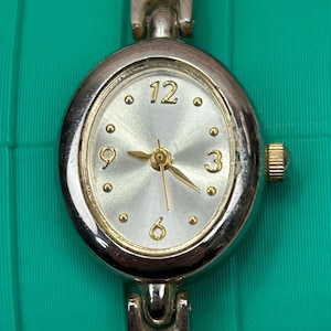 Puede incluir: Reloj de pulsera plateado con esfera ovalada y esfera plateada. El reloj presenta manecillas e indicadores de hora dorados, con los números 12, 3, 6 y 9 claramente visibles. La correa del reloj también es plateada.