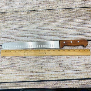 Puede incluir: Un cuchillo de pan de acero inoxidable con mango de madera. El cuchillo tiene una hoja dentada y mide aproximadamente 43 cm de largo. El mango es de color marrón oscuro y tiene tres remaches. Una regla de madera está al lado del cuchillo.