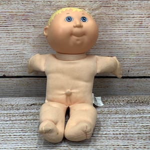 Puede incluir: Una muñeca Cabbage Patch Kid vintage con un cuerpo suave color carne y una cabeza redonda. La muñeca tiene ojos azules, una nariz pequeña y una ligera sonrisa. Su cabello rubio está peinado en un mechón. La muñeca está sentada con los brazos extendidos.