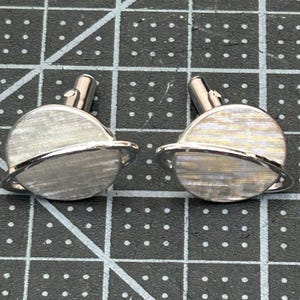 Op de afbeelding: Een paar zilveren manchetknopen met een planeetontwerp. Elke manchetknop heeft een getextureerd, cirkelvormig lichaam met een ring eromheen en een zilveren pin. De manchetknopen worden op een zwart oppervlak weergegeven.