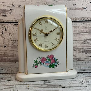 May include: A white ceramic quartz clock with a gold-tone bezel and Roman numerals. The clock face has the letters "PS" and the word "QUARTZ". The clock is decorated with pink and blue floral accents and gold trim.