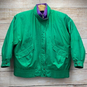 May include: A bright green jacket with a purple collar and lining. The jacket has a zipper closure, two front pockets, and buttoned cuffs. The jacket is hanging on a wooden hanger.