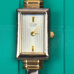 Könnte beinhalten: Eine goldfarbene Citizen Quartz Armbanduhr mit rechteckigem Zifferblatt. Das Zifferblatt ist silberfarben mit goldenen Zeigern und Markierungen. Die Uhr hat eine goldene Krone und ein goldenes Armband. Die Uhr befindet sich vor einem türkisfarbenen Hintergrund.