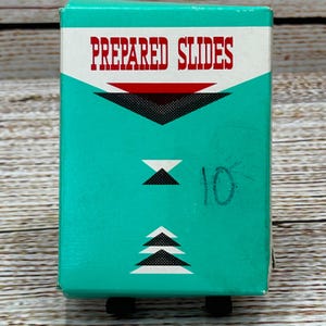 Pode incluir: Uma caixa vintage de lâminas preparadas, com um design turquesa e branco e o texto "PREPARED SLIDES" em vermelho. A caixa tem um padrão geométrico e o número "10" está escrito na lateral. A caixa está sobre uma superfície de madeira.