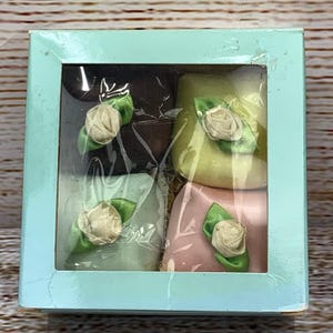 Puede incluir: Una caja cuadrada azul claro con una ventana transparente, que contiene cuatro artículos pequeños envueltos. Cada artículo es de un color diferente: marrón, amarillo, verde claro y rosa. Cada uno está decorado con una pequeña rosa de tela blanca y hojas verdes.