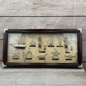 May include: A framed nautical display featuring a small sailboat and various rope knots. The background is a vintage map. The frame is dark brown with a glass cover. The knots are made of beige rope and are labeled with small brass plates.