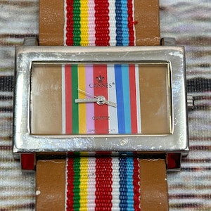 May include: A rectangular silver watch with a brown leather strap and a colorful striped fabric band. The watch face features vertical stripes and the word "Cannes". The watch is marked "Quartz" and "Japan".