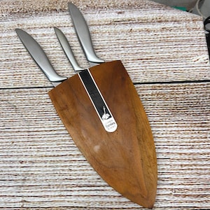 Puede incluir: Un juego de tres cuchillos de acero inoxidable con hojas curvas, guardados en un bloque de cuchillos de madera. El bloque es de color marrón cálido y tiene forma de escudo. Los cuchillos son plateados y el bloque tiene una etiqueta que dice "Legendary Blades".