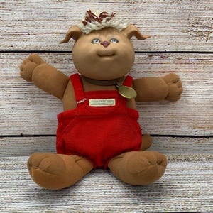 Puede incluir: Una muñeca Koosa de Cabbage Patch Kids vintage. La muñeca tiene una cara bronceada con ojos azules, orejas marrones y un peinado de hilo marrón y blanco. Lleva un peto de pana rojo y un collar marrón con una etiqueta dorada.
