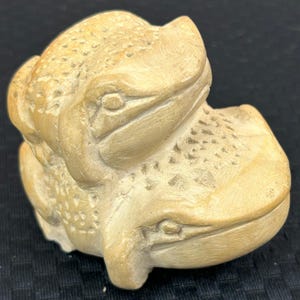 May include: A carved sculpture of two frogs, side by side, in a light beige color. The frogs have detailed features, including textured skin and visible eyes. The sculpture is likely made of stone or a similar material.
