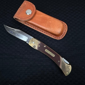 May include: A folding pocket knife with a brown and brass handle, and a silver blade. The knife is next to a brown leather sheath. The blade has the text "J.A.I. SAFETY 92". The handle has the text "OLD TIMER".