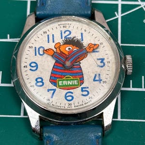 May include: Vintage Bradley watch with Ernie from Sesame Street on the face. The watch has a silver case, a white dial with blue numerals, and a blue leather strap. Ernie is wearing a red and blue striped shirt. The watch is marked "SWISS MADE".