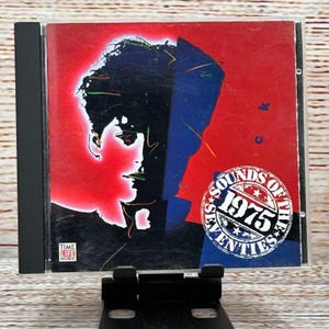 Puede incluir: Una caja de CD con una portada roja y azul que presenta la silueta de un rostro. El texto "SOUNDS OF THE SEVENTIES 1975" está en un diseño circular. El logotipo de Time Life Music está en la esquina inferior izquierda.