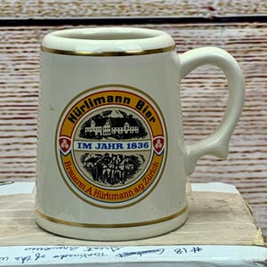 Pode incluir: Um pequeno caneco de cerveja de cerâmica branca com borda e alça douradas. O caneco apresenta um emblema circular com as palavras "Hürlimann Bier" e "IM JAHR 1836". O emblema também inclui uma representação de um edifício e figuras.