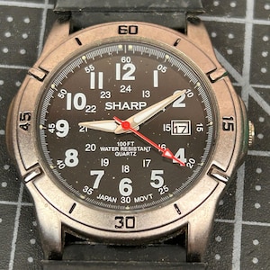 May include: A SHARP quartz watch with a black dial, white numerals, and luminous hands. The watch features a date window at the 3 o'clock position and is water-resistant to 30.5 metres. The watch has a black rubber strap.