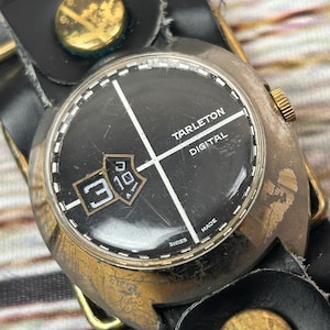 May include: Vintage Tarleton Digital watch with a black face, gold accents, and a black leather band. The watch face displays the date and the words "TARLETON DIGITAL" and "MADE SWISS". The watch has a gold crown and a black leather strap.