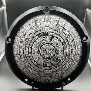 May include: A black, circular metal plate featuring a detailed, etched design of the Aztec calendar. The intricate pattern is in silver against the black background. The plate has several mounting holes around the edge.