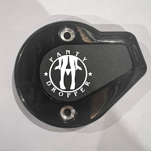 May include: Black, oval-shaped metal object with the text "PANTY DROPPER" in white. The center has a circular design with a silhouette of legs. Two holes are visible for mounting.