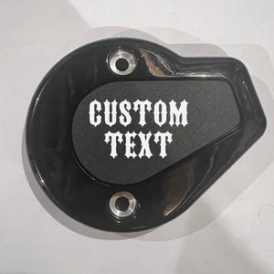May include: Black motorcycle gas cap cover with a white custom text decal that says "CUSTOM TEXT".
