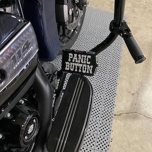 May include: A black motorcycle with a chrome footpeg and a black footboard. The footboard has a black metal plate with the words "Panic Button" in white letters.