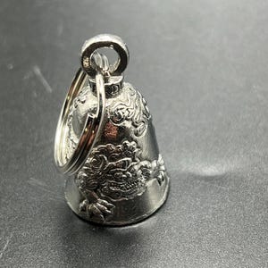 May include: Silver-colored bell with a detailed dragon design. The bell has a loop for attaching to a keychain. The dragon is intricately carved, adding texture and visual interest to the bell's surface. The bell is made of metal and has a shiny finish.