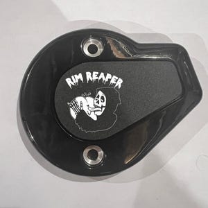 May include: Black motorcycle part with the text "RIM REAPER" and a graphic of the Grim Reaper. The part has a glossy finish and two silver-colored mounting holes.