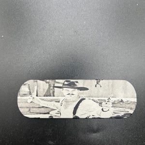May include: A black and white image of a man in a cowboy hat and suspenders, holding a gun. The image is on a small, rectangular object with rounded edges. The background is a blurred outdoor scene.
