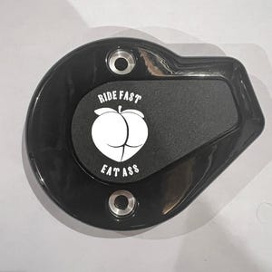 May include: Black motorcycle part with the text "RIDE FAST EAT ASS" and a white graphic of a butt. The part has two holes for mounting and a rounded shape.