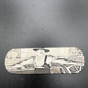 May include: A rectangular, light-colored object with a black and white image of a man in a hat and suspenders. The man has a mustache and is standing with his arms outstretched. The object appears to be made of wood or a similar material.