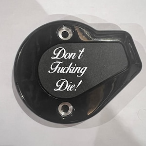 May include: Black motorcycle part with the text "Don't Fucking Die!" in white script. The part has a rounded shape with a rectangular inset and two circular mounting holes.