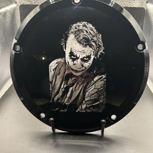 May include: A black, circular motorcycle part featuring a detailed, grayscale image of the Joker. The image is centered on the part, with the Joker's face and upper body visible. The part has several mounting holes around the edge.