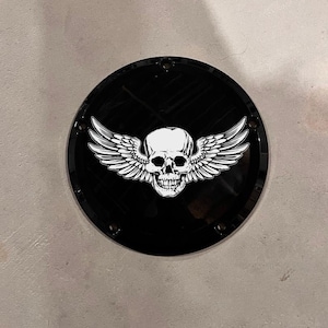 May include: A black, circular emblem featuring a white skull with feathered wings. The skull has detailed facial features. The emblem is secured with four small screws. The design is centered on the black surface.