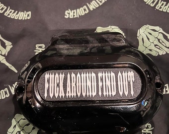 May include: Black motorcycle engine cover with a white sticker that reads "FUCK AROUND FIND OUT".