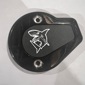 May include: Black, oval-shaped motorcycle part with a textured center and a white shark graphic. The part has two silver mounting holes and a glossy finish. The shark graphic is a detailed illustration with sharp teeth.