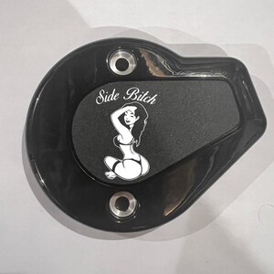 May include: Black motorcycle part with a matte finish. The part has a rounded shape with a rectangular inset. The inset features a white graphic of a woman and the text "Side Bitch". Two silver-colored holes are visible.
