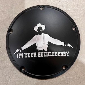 May include: Black round motorcycle cover with a white silhouette of a man wearing a hat and holding two guns. The text "I'M YOUR HUCKLEBERRY" is written below the silhouette.