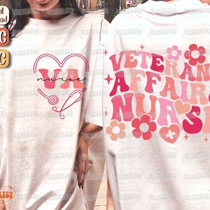 Puede incluir: Dos camisetas blancas con diseños gráficos rosas y rojos. La camiseta delantera tiene un corazón con las letras "VA" dentro y un estetoscopio. La camiseta trasera tiene el texto "Veterans Affairs Nurse" en una fuente retro con corazones y flores.