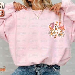 May include: A light pink sweatshirt with a graphic of a ghost with flowers and other objects. The ghost is white with orange and brown accents. The sweatshirt has a relaxed fit and long sleeves. The words "Digital download" and "PNG" are in the upper left corner.