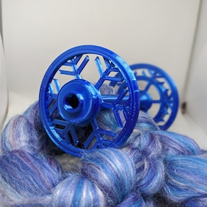 Può includere: Una ruota da filatura blu a forma di fiocco di neve con un gomitolo di lana blu e bianco.