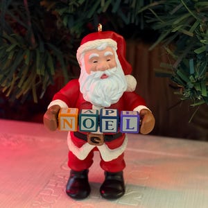 Peut inclure: Une figurine du Père Noël portant un costume rouge avec des bordures blanches, une ceinture noire à boucle dorée et des bottes noires. La figurine tient quatre blocs carrés colorés devant sa poitrine, formant le mot NOEL en grandes lettres.