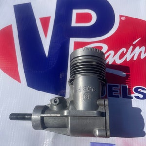 Puede incluir: Un motor de avión modelo de plata con el texto "VECO 61" impreso en el lateral. El motor está sobre una superficie blanca con un logotipo rojo, blanco y azul en el fondo.
