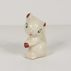 May include: A small white ceramic figurine of a squirrel with red accents on the ears and paws. The squirrel is sitting upright with its tail curled behind it.