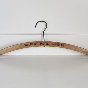 May include: A curved wooden clothes hanger with a dark metal hook. The wood has a natural light brown grain and features the black printed text WALDORF HOTEL, LONDON. centered on the front. The hanger has notched ends for garment straps.