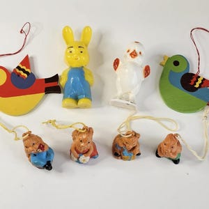 May include: A collection of vintage ornaments featuring a yellow rabbit in blue overalls, a white chick, and colorful bird-shaped ornaments. Small bear figurines with blue and red clothing are also included. Each ornament has a string for hanging.