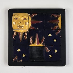 May include: A square black frame containing a grid of nine tiles. The tiles feature a yellow sun face with gold rays, dark blue backgrounds, and small gold stars. Some tiles have raised, pointed, maroon and gold sun ray accents.
