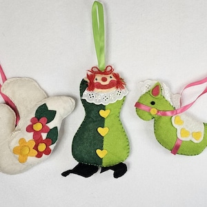 May include: Three handmade felt ornaments: a white bird with floral accents, a green and black clown figure, and a green horse with a pink ribbon. Each ornament has a ribbon for hanging. The ornaments are likely for holiday or seasonal decor.