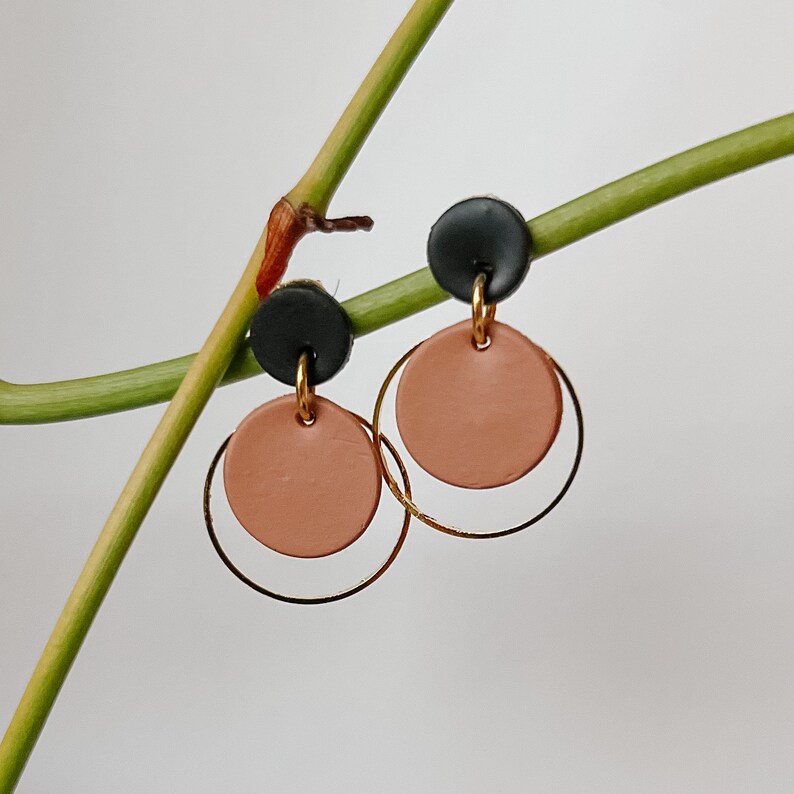 May include: A pair of earrings with a peach colored circle and a gold hoop. The earrings have a black accent on the top.
