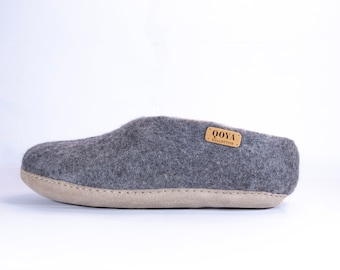 May include: A gray felt slipper with a light tan sole. The slipper has a stitched seam around the sole and a small rectangular tag that reads "OOYA COLLECTIVE". The slipper is designed for indoor wear.