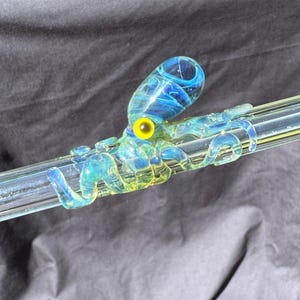 Könnte beinhalten: Eine transparente Glaspfeife mit einem blau-gelben Oktopus-Design. Der Oktopus hat ein gelbes Auge und ist an dem transparenten Glasrohr befestigt. Die Pfeife liegt auf einer schwarzen Oberfläche.